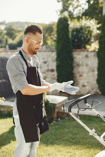 A man wearing a black apron, white gloves, and sunglasses is standing in a well-maintained garden during daylight hours, preparing for gardening work. Behind him, there is a stone wall with a row of t